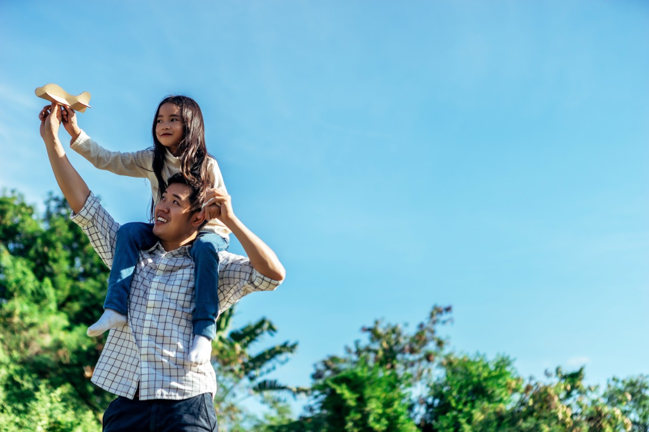 Father with daughter on his shoulders playing with a paper airplane outside on a sunny day